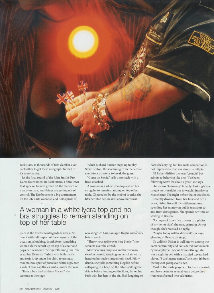 Close-up of a darts player's arm with tattoos and a gold watch, holding three darts with Union Jack flights.