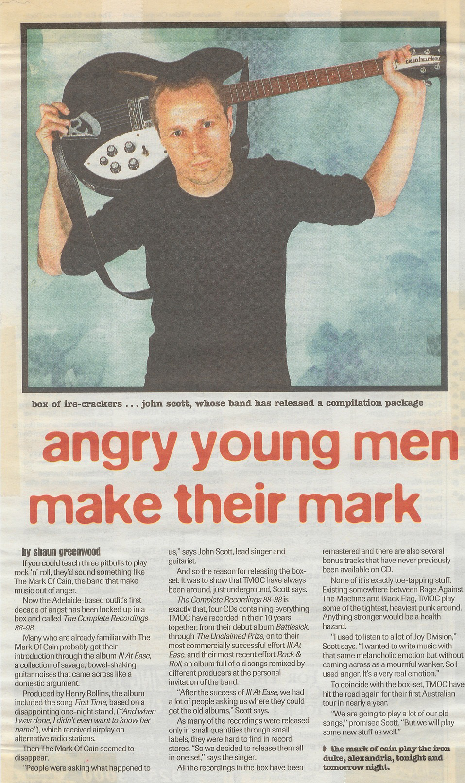 John Scott of The Mark Of Cain holds a black Rickenbacker guitar behind his head over a news article about the band.